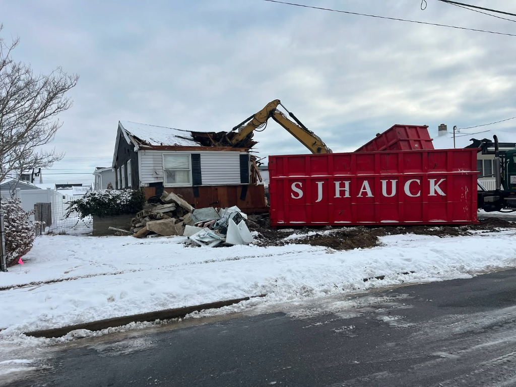SJ Hauck red dumpster in foreground with excavator demolishing a snow-covered house in the background