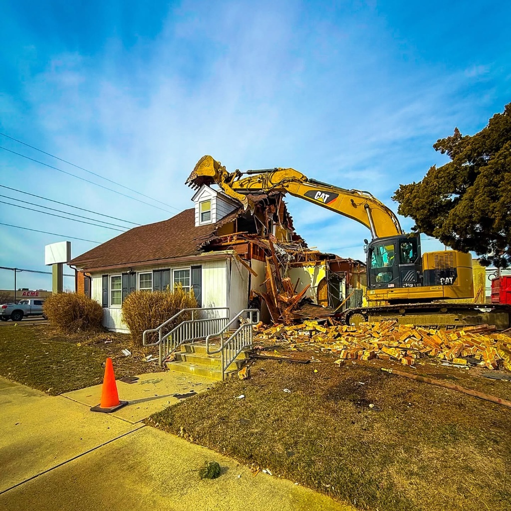 CAT excavator tearing through a brick building during active demolition, debris scattered across the ground, blue sky above