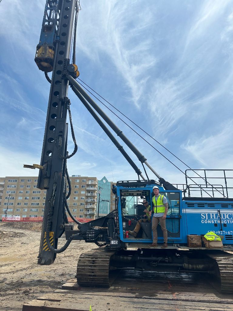 SJ Hauck Construction worker in safety vest standing on a large blue drill rig at an active job site with city buildings in the background